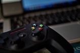 A video game controller with colorful buttons, including green, red, yellow, and blue, placed in front of a blurred laptop keyboard. The focus is on the controller with the surroundings slightly out of focus, indicating a casual gaming setup.