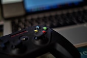 A video game controller with colorful buttons, including green, red, yellow, and blue, placed in front of a blurred laptop keyboard. The focus is on the controller with the surroundings slightly out of focus, indicating a casual gaming setup.