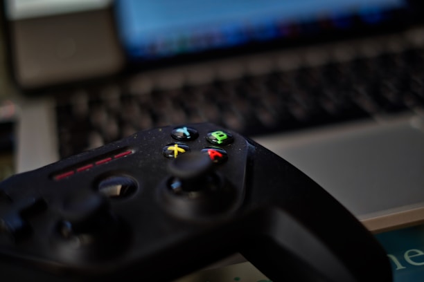 A video game controller with colorful buttons, including green, red, yellow, and blue, placed in front of a blurred laptop keyboard. The focus is on the controller with the surroundings slightly out of focus, indicating a casual gaming setup.