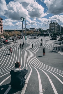 A public space featuring an artistic design of white wavy lines on the dark pavement, creating a striking optical illusion. People are scattered across the area, walking or sitting, with trees and buildings in the background under a partly cloudy sky.