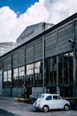 An industrial building with large, rectangular glass windows and concrete walls stands under a sky with fluffy clouds. A small, vintage white car is parked in front of the building, adding a nostalgic element to the scene. The building is labeled 'Refshaleøen.' Wooden planters are positioned along the pavement.