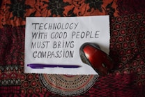 A handwritten note on white paper reads 'Technology with good people must bring compassion.' Below, a purple pen and a red computer mouse are placed on a patterned textile background.