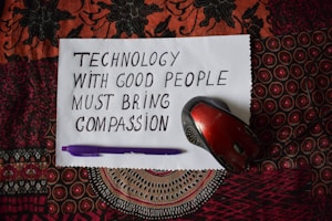 A handwritten note on white paper reads 'Technology with good people must bring compassion.' Below, a purple pen and a red computer mouse are placed on a patterned textile background.