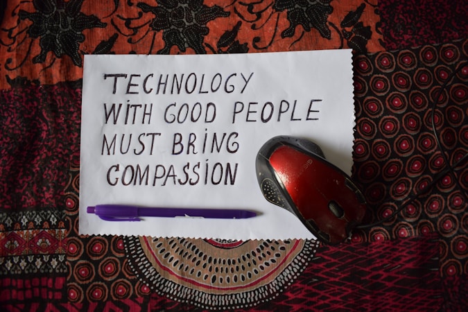 A handwritten note on white paper reads 'Technology with good people must bring compassion.' Below, a purple pen and a red computer mouse are placed on a patterned textile background.