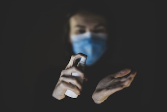 A person wearing a blue mask holds a spray bottle aimed at their other hand, which appears to be opened and ready to receive the spray. The background is dark, highlighting the hands and the spray bottle.