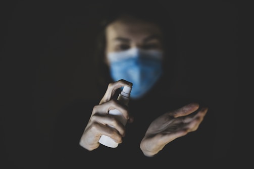 A person wearing a blue mask holds a spray bottle aimed at their other hand, which appears to be opened and ready to receive the spray. The background is dark, highlighting the hands and the spray bottle.