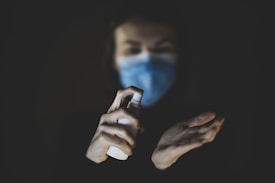 A person wearing a blue mask holds a spray bottle aimed at their other hand, which appears to be opened and ready to receive the spray. The background is dark, highlighting the hands and the spray bottle.