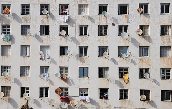 A weathered apartment building facade featuring multiple rows of identical windows, each with a satellite dish attached. Clothes are hung outside the windows, suggesting residents use the space for laundry drying. The walls show significant wear with peeling paint and rust stains, creating an atmosphere of age and urban decay.