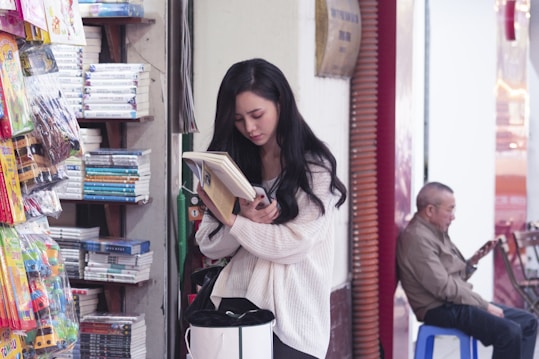 A young woman with long dark hair is engrossed in reading a book while holding a smartphone in her hand. She is standing in a shop filled with stacked books and colorful packaged toys. An older man, seated on a blue stool in the background, is looking at a smartphone.