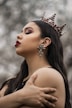 A person with long dark hair is wearing a decorative crown with jewels and intricate details. They have dramatic makeup, including bold red lipstick and eyeshadow. Large star-shaped earrings hang from the ears. Their expression is serene and contemplative as they hold their own shoulders.