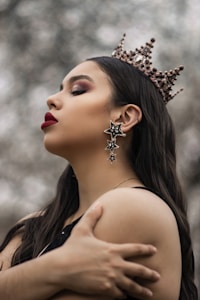 A person with long dark hair is wearing a decorative crown with jewels and intricate details. They have dramatic makeup, including bold red lipstick and eyeshadow. Large star-shaped earrings hang from the ears. Their expression is serene and contemplative as they hold their own shoulders.