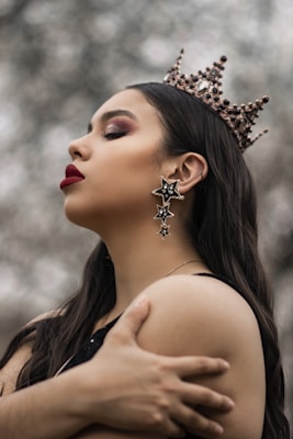 A person with long dark hair is wearing a decorative crown with jewels and intricate details. They have dramatic makeup, including bold red lipstick and eyeshadow. Large star-shaped earrings hang from the ears. Their expression is serene and contemplative as they hold their own shoulders.