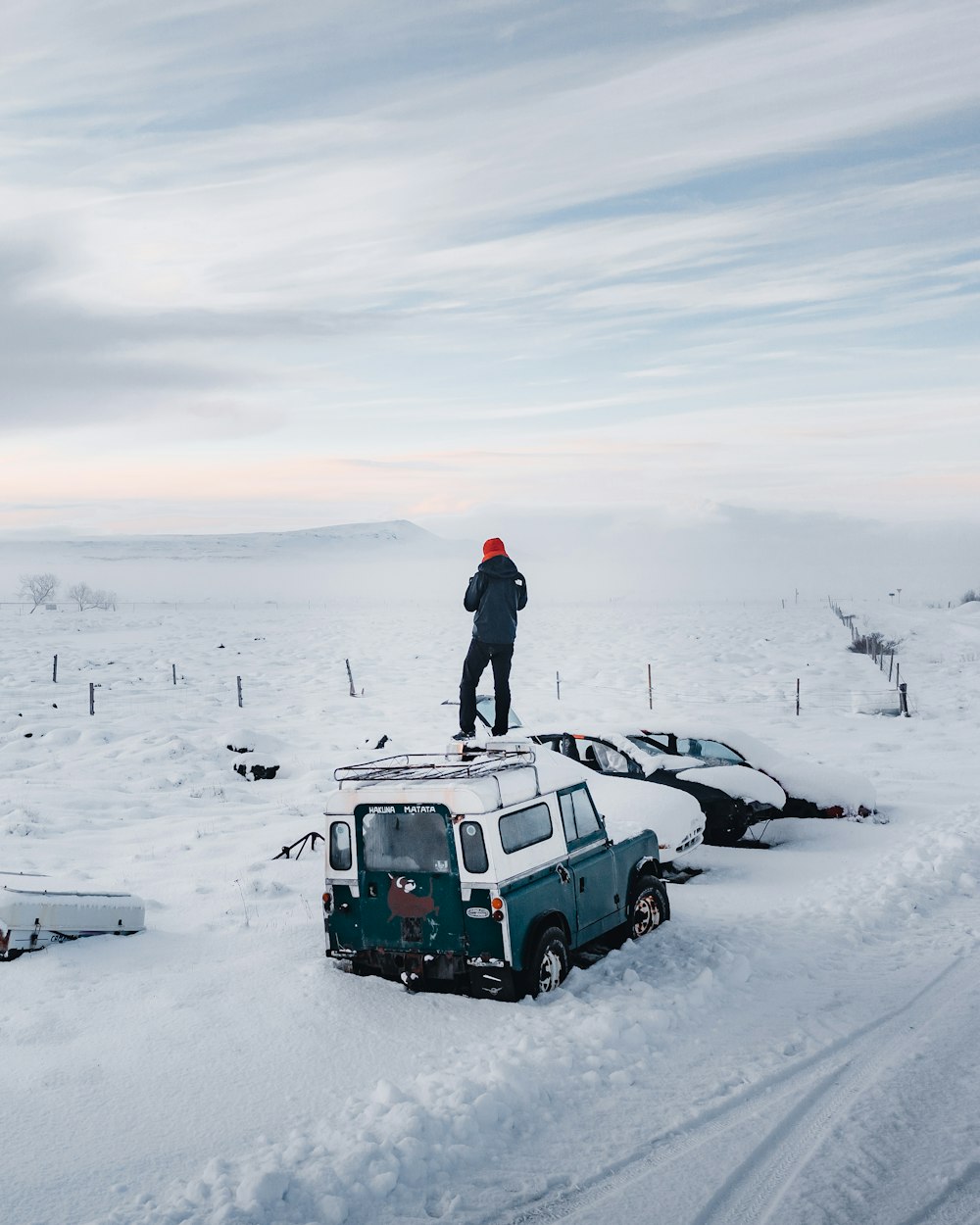 man in black jacket standing on green car on snow covered ground during daytime