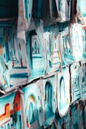 Numerous paintings, each depicting vibrant street scenes with colorful buildings and arched doorways, are displayed side by side. The images are rich in blue and turquoise tones, reflecting a Mediterranean or North African aesthetic.