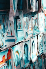Numerous paintings, each depicting vibrant street scenes with colorful buildings and arched doorways, are displayed side by side. The images are rich in blue and turquoise tones, reflecting a Mediterranean or North African aesthetic.