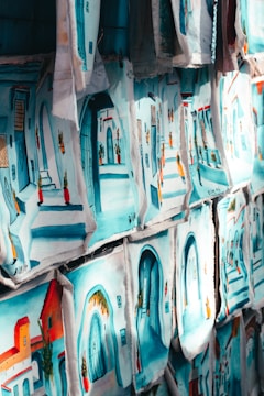Numerous paintings, each depicting vibrant street scenes with colorful buildings and arched doorways, are displayed side by side. The images are rich in blue and turquoise tones, reflecting a Mediterranean or North African aesthetic.