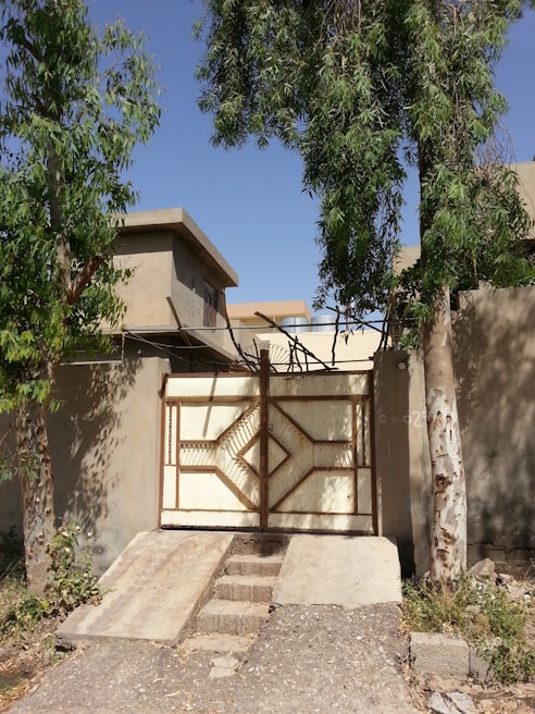 A metal gate with a geometric design stands closed between two tall trees, surrounded by a concrete wall. In the background, a building with flat roofs is partially visible under a clear blue sky. The ground appears dry with some scattered vegetation.