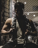A muscular individual wearing a tank top holds a heavy metal chain draped around their shoulders, set against a dimly lit background with a wire fence and brick wall. The atmosphere is shadowy, emphasizing the subject's strength and determination.
