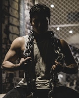 A muscular individual wearing a tank top holds a heavy metal chain draped around their shoulders, set against a dimly lit background with a wire fence and brick wall. The atmosphere is shadowy, emphasizing the subject's strength and determination.