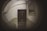 A fisheye view captures an indoor hallway with a closed door at the center. The walls are painted in two tones, a light grey above and a muted green below. An old-fashioned staircase railing is visible on the left side, and a window with horizontal bars is positioned on the right wall. The image has a distortion effect typical of fisheye lenses.