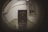 A fisheye view captures an indoor hallway with a closed door at the center. The walls are painted in two tones, a light grey above and a muted green below. An old-fashioned staircase railing is visible on the left side, and a window with horizontal bars is positioned on the right wall. The image has a distortion effect typical of fisheye lenses.