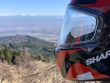 A motorcycle helmet with a glossy finish is positioned in the foreground, overlooking a vast and clear mountainous landscape. The view includes a mix of green forests, distant mountains, and a blue sky with a few clouds.