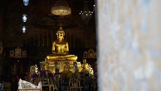 A large golden Buddha statue is prominently displayed in an ornate interior setting. The statue is surrounded by smaller statues, intricate decorations, and floral arrangements. The lighting is dim, creating an atmosphere of reverence and tranquility. The image includes hanging lamps and a chandelier that add to the ambiance.