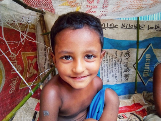 A young child with short dark hair and a slight smile is inside a makeshift shelter. The shelter is constructed from colorful woven materials and bamboo sticks. The child is wrapped in a bright blue cloth and surrounded by bags with text in a foreign script.