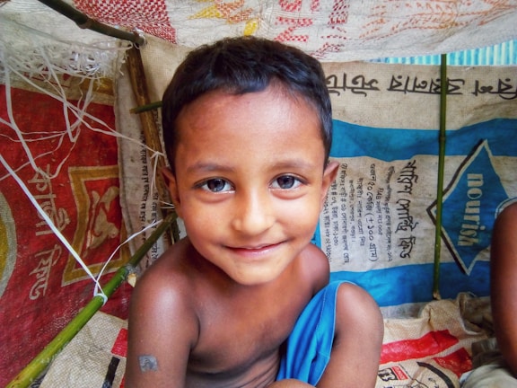 A young child with short dark hair and a slight smile is inside a makeshift shelter. The shelter is constructed from colorful woven materials and bamboo sticks. The child is wrapped in a bright blue cloth and surrounded by bags with text in a foreign script.
