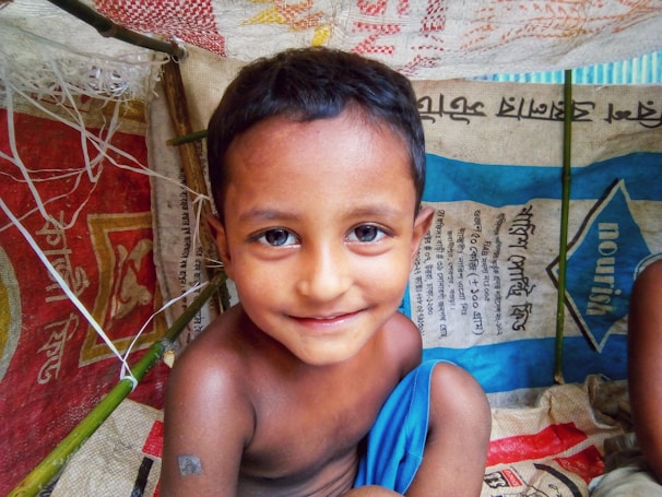 A young child with short dark hair and a slight smile is inside a makeshift shelter. The shelter is constructed from colorful woven materials and bamboo sticks. The child is wrapped in a bright blue cloth and surrounded by bags with text in a foreign script.