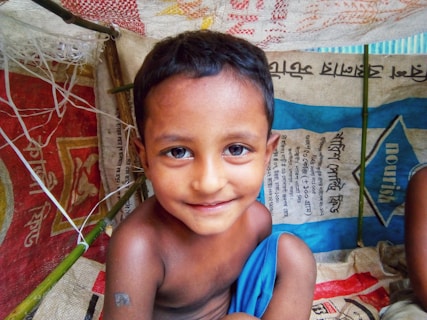 A young child with short dark hair and a slight smile is inside a makeshift shelter. The shelter is constructed from colorful woven materials and bamboo sticks. The child is wrapped in a bright blue cloth and surrounded by bags with text in a foreign script.
