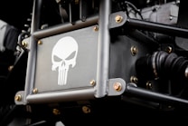 A metal panel featuring a stylized skull logo reminiscent of the Punisher emblem, surrounded by metal rods and bolts. The setting appears to be part of a vehicle or machinery with visible mechanical components.