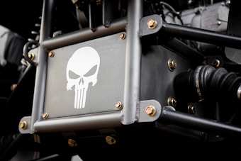 A metal panel featuring a stylized skull logo reminiscent of the Punisher emblem, surrounded by metal rods and bolts. The setting appears to be part of a vehicle or machinery with visible mechanical components.