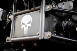 A metal panel featuring a stylized skull logo reminiscent of the Punisher emblem, surrounded by metal rods and bolts. The setting appears to be part of a vehicle or machinery with visible mechanical components.