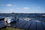 Two scuba diving tanks are placed on a wooden dock beside a body of water with waves. The sky is clear with several fluffy white clouds, and the surroundings hint at a natural setting with distant trees near the horizon.