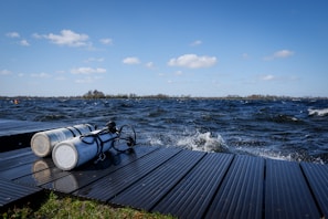 Two scuba diving tanks are placed on a wooden dock beside a body of water with waves. The sky is clear with several fluffy white clouds, and the surroundings hint at a natural setting with distant trees near the horizon.