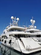 A luxurious white yacht docked at the marina under a clear blue sky. The yacht features multiple decks, satellite antennas, and is securely tied to the dock.