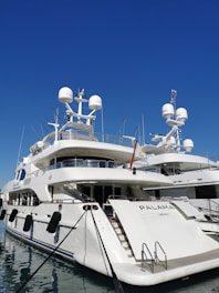 A luxurious white yacht docked at the marina under a clear blue sky. The yacht features multiple decks, satellite antennas, and is securely tied to the dock.