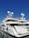A luxurious white yacht docked at the marina under a clear blue sky. The yacht features multiple decks, satellite antennas, and is securely tied to the dock.