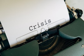 A close-up view of a typewriter with a sheet of paper displaying the word 'Crisis' in bold, black letters. The typewriter has a dark green casing.