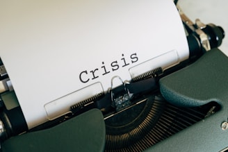 A close-up view of a typewriter with a sheet of paper displaying the word 'Crisis' in bold, black letters. The typewriter has a dark green casing.