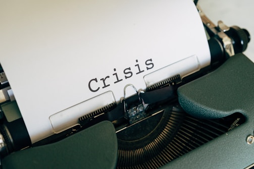 A close-up view of a typewriter with a sheet of paper displaying the word 'Crisis' in bold, black letters. The typewriter has a dark green casing.