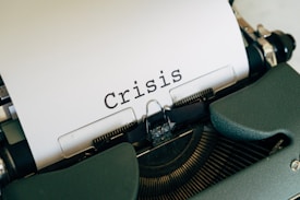 A close-up view of a typewriter with a sheet of paper displaying the word 'Crisis' in bold, black letters. The typewriter has a dark green casing.