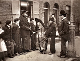 A group of boys and a man gathered around a street musician playing a harp. The scene is set on a sidewalk outside a row of brick houses with windows and shutters. The boys are dressed in period clothing, including hats and jackets, standing in various poses of attention and interest. The musician appears focused on playing, creating a focal point for the gathering.