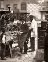 A black and white vintage photograph of a street scene featuring a man wearing a hat and apron working with tools at a market stall. He is surrounded by various tools laid out on the table. Another man, also wearing a hat and light coat, stands nearby, and a child stands in front of the stall. In the background are blurred outlines of buildings and a wagon.