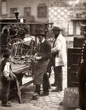 A black and white vintage photograph of a street scene featuring a man wearing a hat and apron working with tools at a market stall. He is surrounded by various tools laid out on the table. Another man, also wearing a hat and light coat, stands nearby, and a child stands in front of the stall. In the background are blurred outlines of buildings and a wagon.