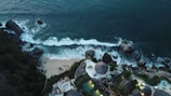 Aerial view of a coastal area with waves crashing onto the shore, bordered by rocky cliffs and lush greenery. A resort with circular structures and pools is situated near the water's edge, surrounded by tropical vegetation.