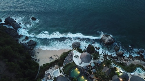 Aerial view of a coastal area with waves crashing onto the shore, bordered by rocky cliffs and lush greenery. A resort with circular structures and pools is situated near the water's edge, surrounded by tropical vegetation.