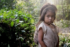 A young child stands in a lush, green forest. The child wears a simple, worn-out garment and has an expression that suggests contemplation or curiosity. The sunlight filters softly through the trees, creating a serene and intimate atmosphere.