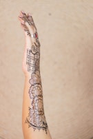 An intricately designed henna or mehndi pattern adorns a person's arm and hand, featuring floral and geometric motifs. The person is wearing a ring with a red gemstone on their finger.